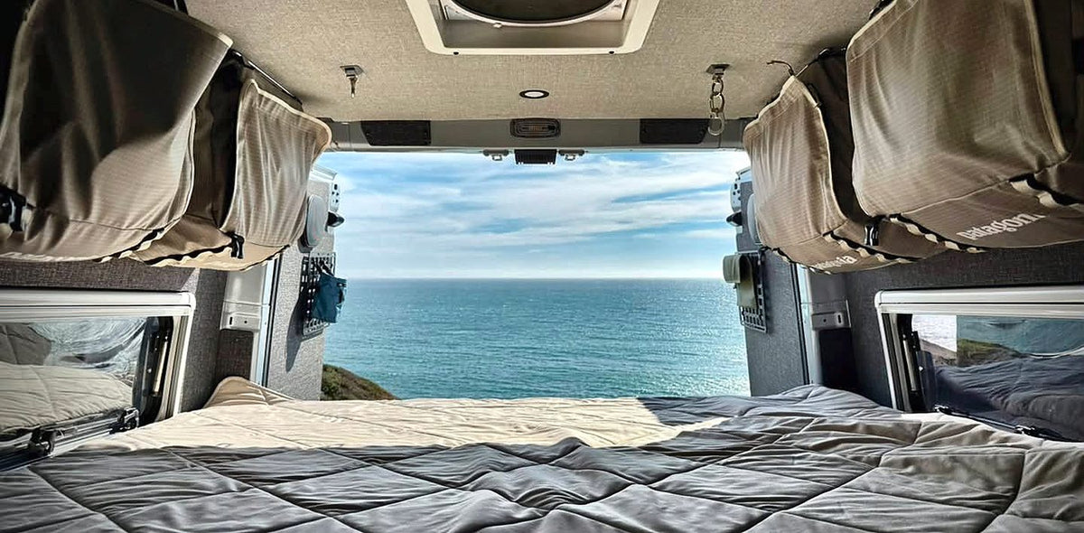 View out the back of a Sprinter van to the ocean, with duffels hanging as cabinets on both sides over the bed